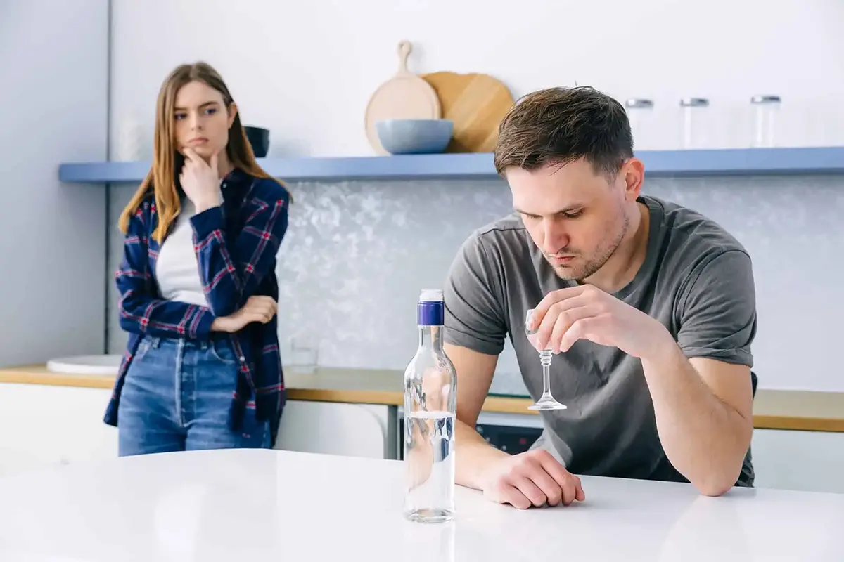 Man drinking in kitchen with wife watching, illustrating how alcohol consumption worsens mental health